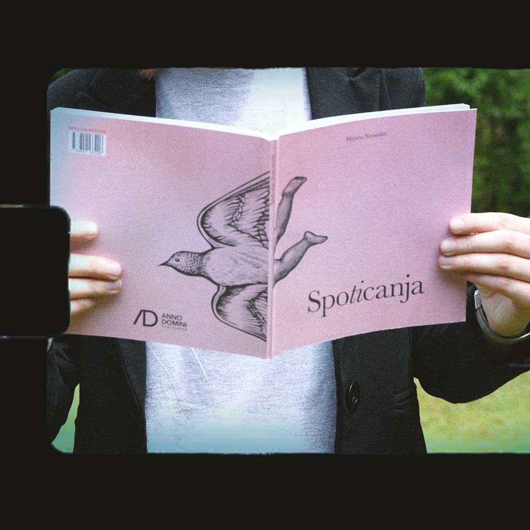 Osoba u crnom sakou i beloj košulji pred licem drži ružičastu knjigu „Spotičanja“. Na koricama knjige je ilustracija ptice sa ljudskom nogom. Lice osobe se ne vidi.